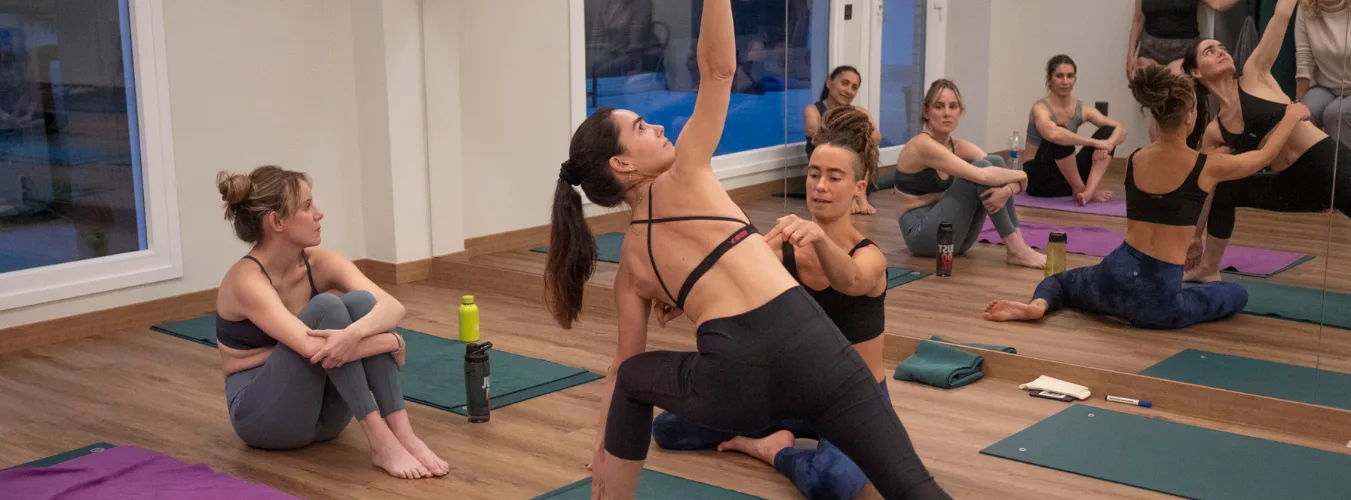 A yoga instructor provides a hands-on adjustment to a student in a side-angle pose while other students sit on their mats and observe in a mirrored studio.