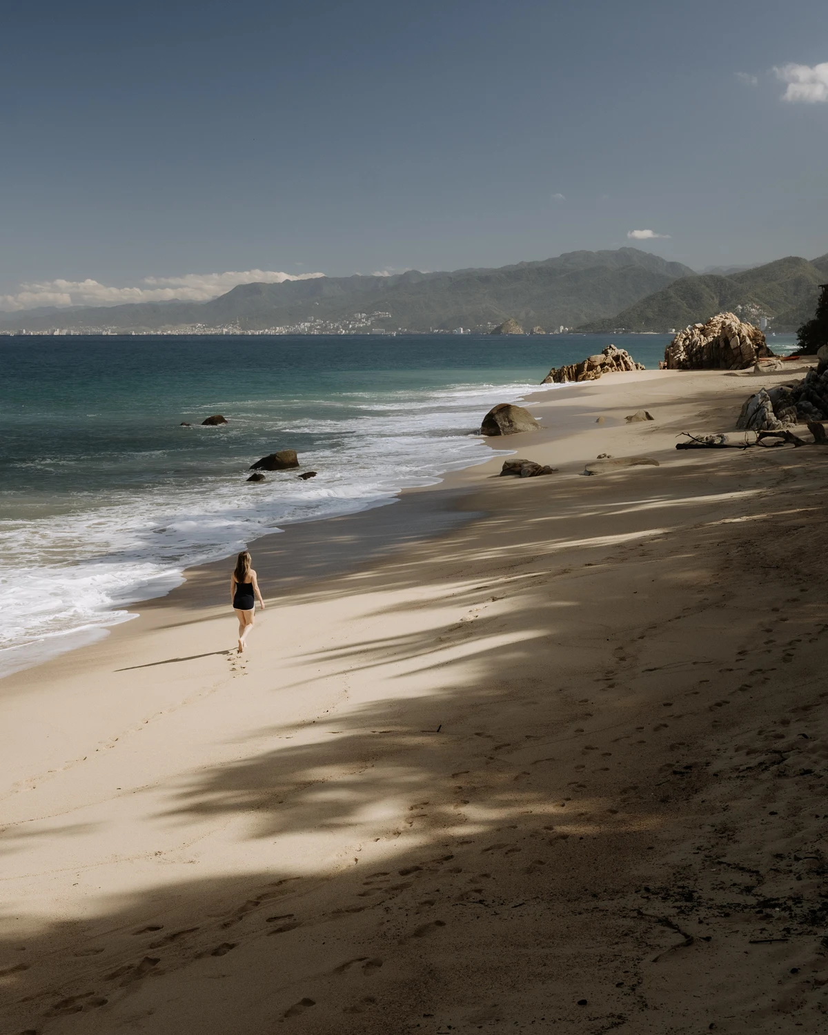 A solitary woman walks barefoot on a wide, sandy beach in Mexico, leaving footprints near the ocean's edge. Dramatic mountain ranges and the distant city of Puerto Vallarta frame the turquoise bay under a clear sky.