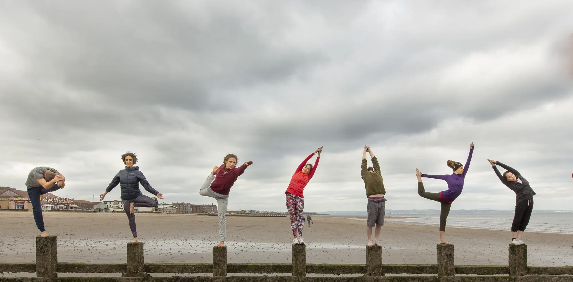 A group of seven yoga practitioners balancing in various postures on wooden posts along a misty, gray coastline during a coastal yoga journey.