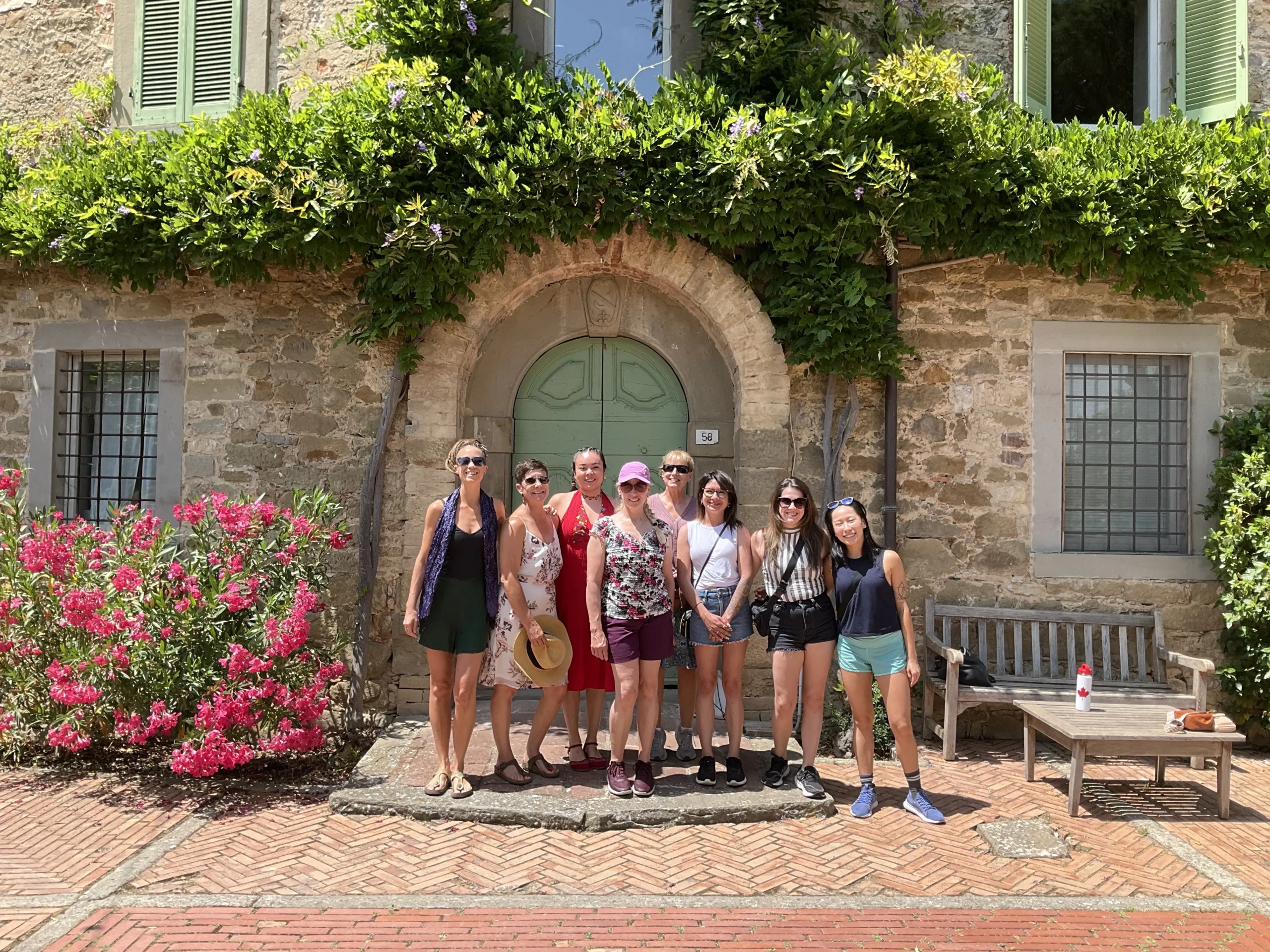 A group of eight women posing for a portrait in front of a historic stone villa surrounded by vibrant pink bougainvillea. 