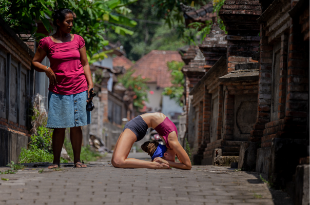 A yoga student practicing a deep kneeling backbend (Kapotasana) on a stone path in a traditional village, representing the immersive "yoga all day" experience of a yoga holiday.