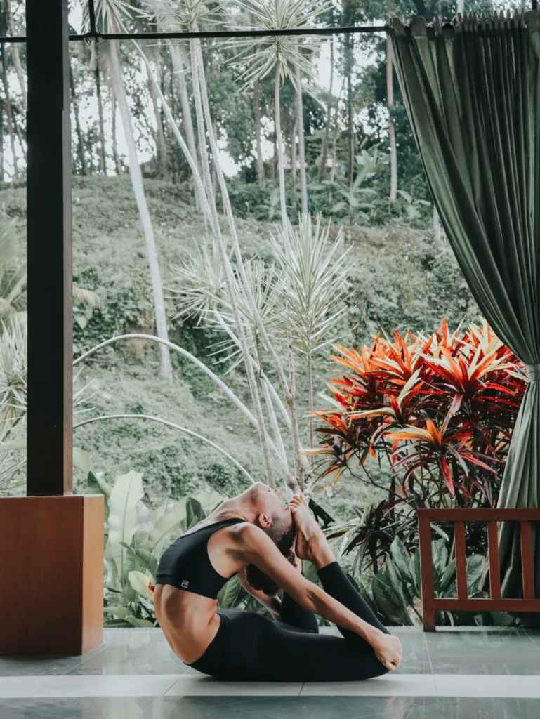 Woman performing a deep backbend yoga pose on a balcony surrounded by tropical plants and greenery