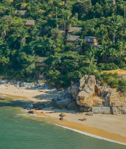 Aerial view of thatched-roof huts and palm trees nestled on a rocky jungle cliffside overlooking a secluded tropical beach.