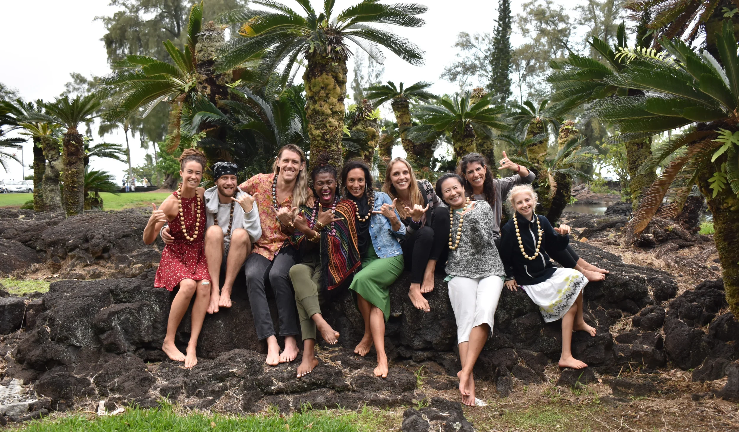 A diverse group of nine smiling e84 Yoga staff members and students sit together on dark volcanic rocks in a tropical setting, wearing leis and making the shaka "hang loose" hand gesture. This highlights the warm, community-focused spirit of Esak Garcia’s teacher training team.